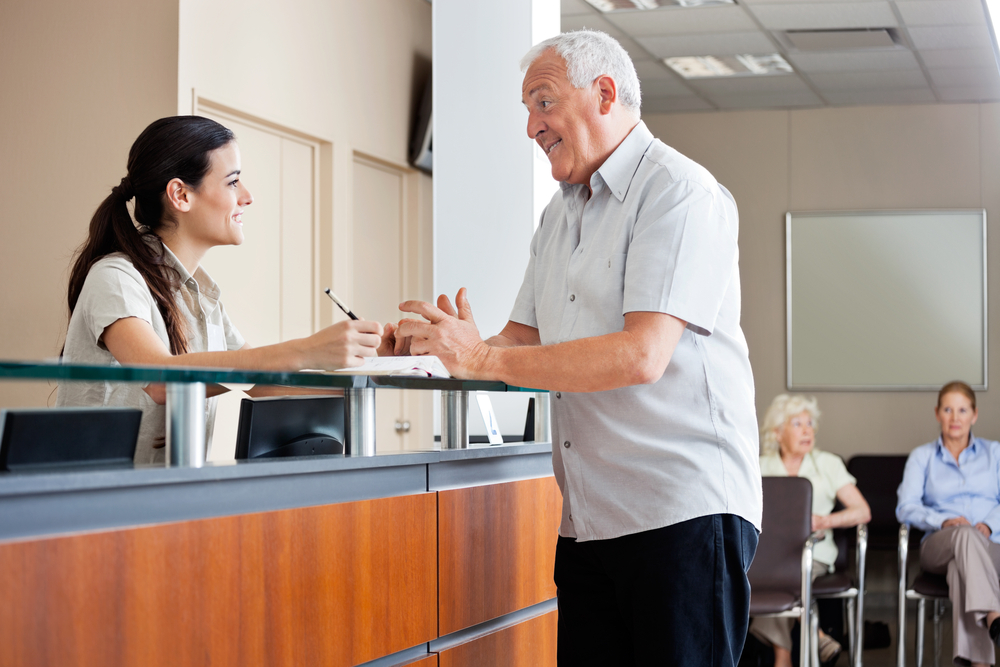 female nurse behind reception desk talking to senior male patient